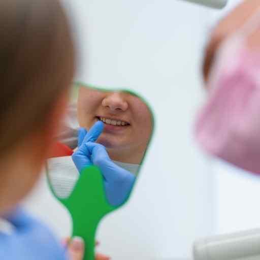 A person holds a green hand mirror reflecting their smile as a dental gloved hand checks their teeth during a dental check-up.
